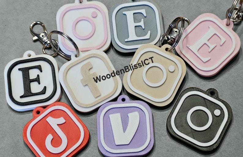 Pu&ograve; includere: Un set di nove portachiavi con diversi loghi di social media e lettere. I portachiavi sono realizzati in diversi colori, tra cui rosa, grigio, beige, nero, bianco, rosso e viola. I portachiavi sono di forma quadrata con angoli arrotondati e hanno un bordo bianco attorno al logo o alla lettera. I portachiavi sono attaccati a anelli portachiavi argentati. Il testo "WoodenBlissICT" &egrave; visibile su uno dei portachiavi.