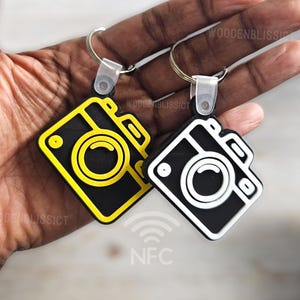 May include: Two camera-shaped keychains with black and white designs. The keychain on the left has a yellow outline and the keychain on the right has a white outline. Both keychains have the word "NFC" printed on them.