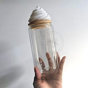 Peut inclure: Un bocal en verre transparent avec un couvercle en bois et un dessus blanc texturé en forme de cône. Le bocal est tenu par une main sur un fond blanc. Le bocal a une forme cylindrique et une large ouverture.