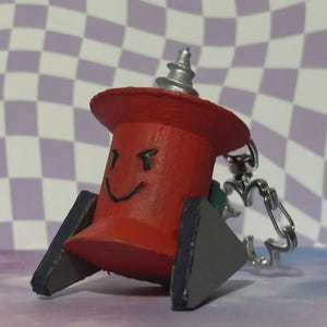 May include: A red, cartoon-like pushpin keychain with a smiling face, black eyebrows, and a silver top. The pushpin has gray and green accents and is attached to a silver keychain.