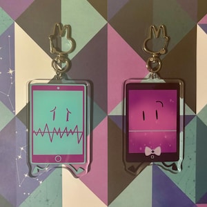 May include: Two clear acrylic keychains with cartoon tablet designs. One has a teal screen with a pink border, featuring two upward arrows and a waveform. The other has a purple screen with a pink border, with two dashes and a bow tie.
