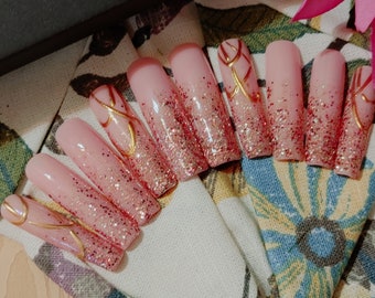Peut inclure: Longues ongles en gel press-on avec un dégradé de paillettes roses et rouges et des accents dorés. Les ongles sont disposés en demi-cercle sur un fond de tissu floral.