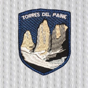 以下が含まれることがあります： 「TORRES DEL PAINE」の文字と山脈が刺繍されたパッチ。パッチは青い縁取りと盾の形をしています。山はベージュとグレーで、濃い青色の空を背景にしています。