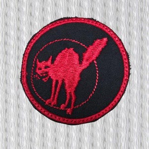 May include: Embroidered patch featuring a red cat silhouette within a black circle, outlined with red stitching. The cat is arched, with its tail raised. The patch has a red stitched border and is set against a textured white background.