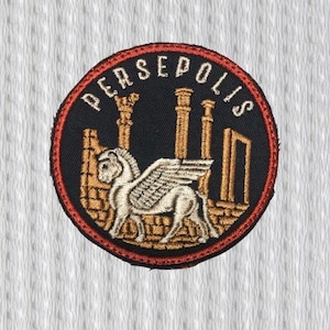 Może przedstawiać: Okrągła, haftowana naszywka z napisem "PERSEPOLIS" w kolorze białym. Naszywka ma czarne tło z uskrzydlonym koniem i elementami architektonicznymi w kolorze beżowym i złotym, obramowana czerwoną obwódką.
