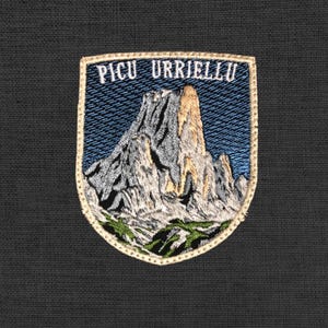 Könnte beinhalten: Gestickter Aufnäher mit einer Berglandschaft und dem Text "PICU URRIELLU". Der Aufnäher hat einen blauen Hintergrund mit einem detaillierten Bergdesign in Grau-, Braun- und Grüntönen, umrahmt von einem beigen Rand.
