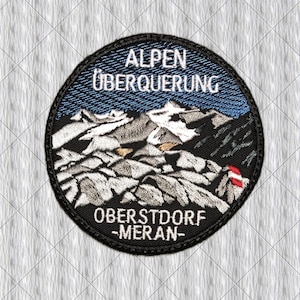 Op de afbeelding: Een ronde geborduurde patch met een berglandschap met besneeuwde toppen. De tekst ALPEN ÜBERQUERUNG staat bovenaan en OBERSTDORF -MERAN- onderaan. Het ontwerp bevat een klein rood-wit vlagdetail aan de rechterkant van de afbeelding.