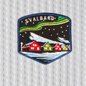 May include: Embroidered patch depicting a winter scene with the word "SVALBARD". The design shows red and green houses under a starry night sky with an aurora borealis. The patch has a blue border.