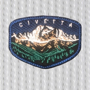 May include: Embroidered patch featuring a mountain landscape with the word "CIVETTA" across the top. The patch has a dark blue border and a black background. The mountain is depicted in white and gold, with green trees at the base.