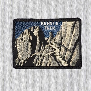 May include: Embroidered patch featuring a mountain range with a hiking trail. The patch has a black border and the text "BRENTA TREK" in white. The background is blue, and the mountains are beige and black.