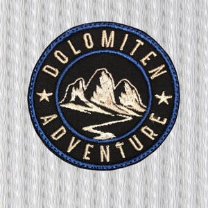 May include: A circular patch with the words "DOLOMITEN ADVENTURE" embroidered in white on a black background. The patch features a blue border and a mountain range design in white and beige.
