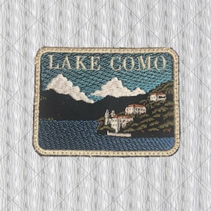 Könnte beinhalten: Gestickter Aufnäher mit einer Landschaft des Comer Sees. Der Aufnäher zeigt den Text "LAKE COMO" über einer Landschaft mit blauem See, weißen Wolken und grünen Hügeln mit Gebäuden. Der Aufnäher hat einen goldenen Rand.