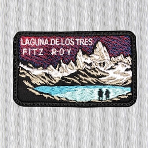 Puede incluir: Parche bordado con el texto "LAGUNA DE LOS TRES FITZ ROY". Representa un paisaje montañoso, un lago azul y un cielo colorido. El parche tiene un borde negro y es de forma rectangular.