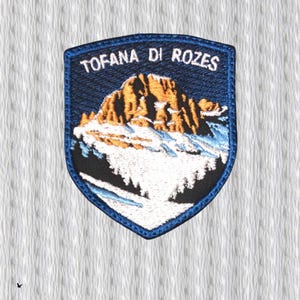 May include: Embroidered patch with the text "TOFANA DI ROZES" above a mountain scene. The patch has a dark blue border and background, with a detailed depiction of a mountain range in shades of brown, white, and blue. The patch is shield-shaped.