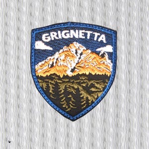 Op de afbeelding: Geborduurde patch met een berglandschap en het woord "GRIGNETTA" bovenaan. De patch heeft een blauwe rand en toont een bergketen met gouden en witte toppen boven een bos.