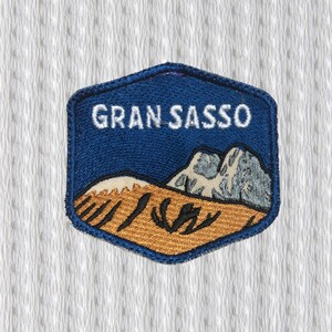 Può includere: Toppa ricamata con bordo esagonale blu e la scritta "GRAN SASSO" in bianco. La toppa presenta un paesaggio con montagne e un primo piano marrone dorato, adatta per giacche o borse.