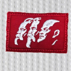 Puede incluir: Un parche rectangular rojo con un diseño bordado en blanco de tres caras estilizadas. El parche tiene un borde cosido rojo y está sobre un fondo blanco texturizado. Las caras están de perfil.