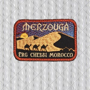 May include: A rectangular embroidered patch featuring a desert landscape with golden sand dunes, a yellow sun, and three black camel silhouettes. The text MERZOUGA is at the top, and ERG CHEBBI MOROCCO is at the bottom, all bordered by a red frame.