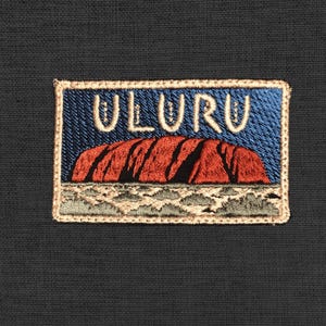 Peut inclure: Écusson brodé avec le mot "ULURU" au-dessus d'une représentation d'Uluru, une grande formation rocheuse de grès en Australie. L'écusson a un fond bleu avec une formation rocheuse rouge et une bordure beige. L'écusson est rectangulaire.
