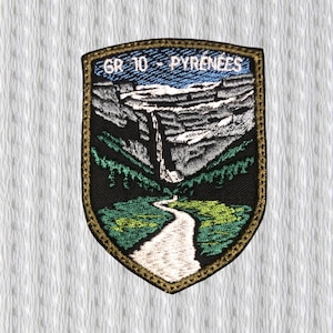 Puede incluir: Parche bordado con forma de escudo que representa un paisaje montañoso con una cascada y un sendero. El texto "GR 10 - PYRENEES" está en la parte superior. El parche tiene un fondo negro con detalles verdes, blancos y azules y un borde dorado.