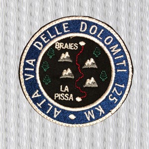 May include: Embroidered patch with a circular design featuring the text "VIA DELLE DOLOMITI" in blue, surrounding a black center. The center depicts a map with mountains, trees, and a red path. The text "ALTA 125 KM" is also included.