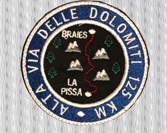 Może przedstawiać: Okrągła, haftowana naszywka z niebieską obwódką i napisem "VIA DELLE DOLOMITI". Czarny środek przedstawia mapę z górami, drzewami i czerwoną ścieżką. Zawiera również tekst "ALTA 125 KM". Dla turystów.