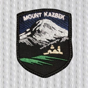 May include: Embroidered patch featuring Mount Kazbek. The patch has a black border and a shield shape. The design includes a mountain, text, and green and tan elements. The text "MOUNT KAZBEK" is embroidered in white.