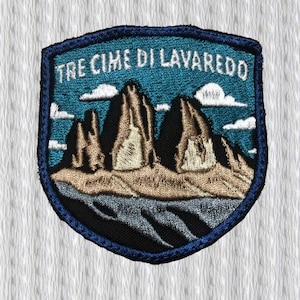 Può includere: Toppa ricamata raffigurante un paesaggio montano con la scritta "TRE CIME DI LAVAREDO" in bianco. La toppa ha un bordo blu e una forma a scudo. Le montagne sono in tonalità marroni e nere, con cielo blu e nuvole bianche.