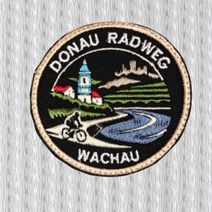 Pode incluir: Patch bordado circular com o texto "DONAU RADWEG WACHAU". O design apresenta um ciclista, um rio, uma igreja e um castelo. O patch é preto com detalhes em bege e branco.