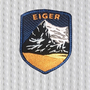 Puede incluir: Parche bordado con un paisaje de montaña y la palabra "EIGER" en naranja. El parche tiene forma de escudo con un borde azul. La montaña es negra y blanca, sobre una base naranja.