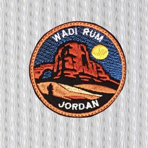 Op de afbeelding: Geborduurde patch met een woestijnlandschap en de tekst "WADI RUM JORDAN". De patch heeft een cirkelvormig ontwerp met een blauwe lucht, een gele zon en een rode rotsformatie. De patch heeft een bruine en oranje rand.