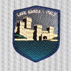 Könnte beinhalten: Gestickter Aufnäher in Schildform mit der Aufschrift "LAKE GARDA - ITALY". Das Design zeigt eine Burgszene über einem blauen See mit einem stürmischen Himmel. Der Aufnäher hat einen blauen Rand.