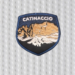 May include: Embroidered patch featuring the word "CATINACCIO" above a mountain range design. The patch has a black base with a blue border and a blue background for the text. The mountain range is depicted in shades of gold and white.