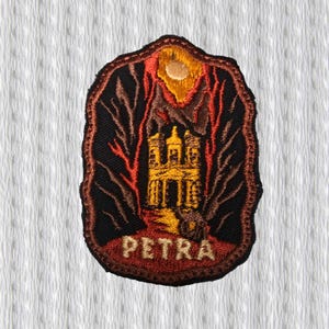 Op de afbeelding: Geborduurde patch met een gedetailleerde illustratie van Petra, Jordanië. Het ontwerp toont een gouden gebouw, bergen en een zon tegen een zwarte achtergrond. Het woord "PETRA" is onderaan in beige geborduurd.