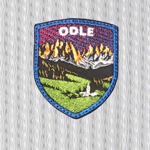 May include: Embroidered patch with a shield shape, featuring a mountain landscape with the word "ODLE" at the top. The patch has a blue border and depicts green fields, brown mountains, and a red and purple sky.