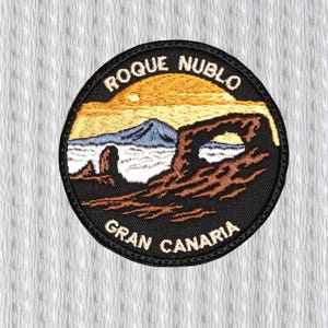 Könnte beinhalten: Runder, gestickter Aufnäher mit dem Text "ROQUE NUBLO" und "GRAN CANARIA". Das Design zeigt eine Berglandschaft mit Sonnenuntergang in Orange und Gelb sowie Felsformationen, alles vor schwarzem Hintergrund.