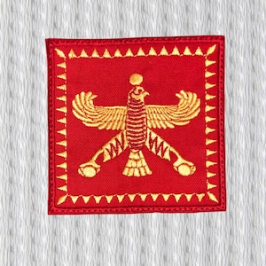 Puede incluir: Parche cuadrado rojo bordado con un pájaro dorado con las alas extendidas y un emblema en forma de sol sobre su cabeza. El parche tiene un borde dorado con detalles triangulares. El diseño está centrado sobre un fondo blanco.