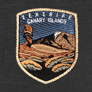 Può includere: Toppa ricamata a forma di scudo con bordo dorato. La toppa presenta la scritta "TENERIFE CANARY ISLANDS" sopra un paesaggio. La scena comprende montagne, formazioni rocciose e un cielo blu.