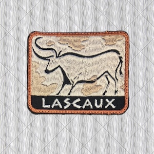 Può includere: Una toppa ricamata rettangolare con un'illustrazione di un toro beige e nero su sfondo testurizzato. La parola LASCAUX è ricamata in lettere maiuscole bianche su una fascia orizzontale nera nella parte inferiore, con un bordo color rame.
