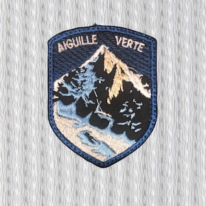 Könnte beinhalten: Ein marineblauer, gestickter Aufnäher mit den Worten "AIGUILLE VERTE" über einer Berglandschaft. Der Berg ist schwarz mit beigen und blauen Akzenten. Der Aufnäher hat einen dunkelblauen Rand und ist schildförmig.