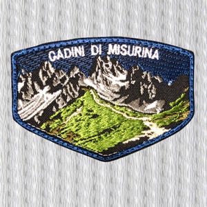 May include: Embroidered patch with a mountain landscape design. The patch features the text "CADINI DI MISURINA" in white letters. The patch has a blue border and depicts green hills and snow-capped mountains.