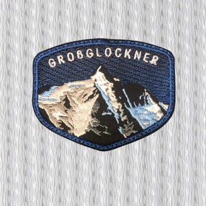 Op de afbeelding: Geborduurde patch met een berglandschap en de tekst "GROBGLOCKNER" in het wit. De patch heeft een donkerblauwe rand en een gestructureerde achtergrond, met de berg in zwart, wit en blauw.