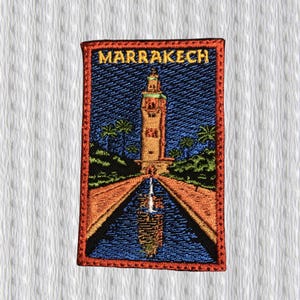 Op de afbeelding: Geborduurde patch met een gedetailleerde illustratie van een bezienswaardigheid in Marrakech. De rechthoekige patch heeft een rode rand en toont een hoge toren, palmbomen en een reflecterend zwembad. Het woord "MARRAKECH" staat bovenaan geborduurd.