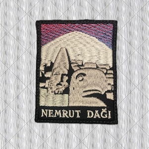 May include: A rectangular embroidered patch featuring a beige depiction of the stone heads at Nemrut Dağı against a dark purple and blue sky. The text NEMRUT DAĞI is stitched in white capital letters at the bottom of the black bordered patch.