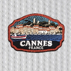 May include: An embroidered patch featuring a coastal scene of Cannes, France. The design includes a red-bordered shape, a blue sea with a small boat, a row of beige buildings, and a dark sky. The text CANNES FRANCE is stitched in white letters.