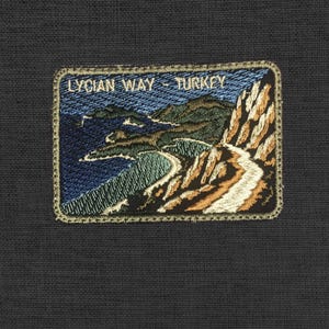 Op de afbeelding: Geborduurde patch met een kustlandschap met de tekst "LYCIAN WAY - TURKEY". De patch toont een kronkelende weg langs een blauwe zee, met groene en bruine kliffen. De patch heeft een gouden rand en is geplaatst tegen een donkere achtergrond.