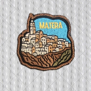 Könnte beinhalten: Ein gestickter Aufnäher mit einer stilisierten Darstellung der Stadt Matera mit beigen Gebäuden, einem hohen Turm und einem hellblauen Himmel. Das Wort MATERA ist in gelben Buchstaben oben eingestickt. Der Aufnäher hat einen dunkelbraunen Rand.