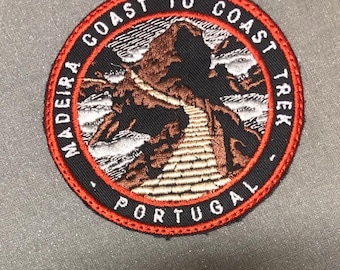 Może przedstawiać: Okrągła naszywka z napisem "MADEIRA COAST TO COAST TREK PORTUGAL" w białych literach na czarnym tle. Naszywka przedstawia brązową scenerię górską z białą ścieżką, obramowaną czerwoną obwódką.