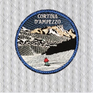 May include: A circular embroidered patch featuring a mountain landscape with snow-capped peaks and a small figure in a red jacket standing on a white slope. The text CORTINA D'AMPEZZO is stitched in white at the top, all bordered by a blue edge.