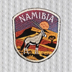 Puede incluir: Un parche bordado en forma de escudo con la palabra NAMIBIA en letras mayúsculas blancas en la parte superior. El diseño muestra un órix de pie en un paisaje desértico con dunas naranjas, la silueta oscura de un árbol y un gran sol naranja.